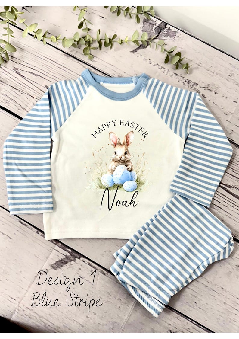 May include: A white long-sleeved shirt with blue and white striped sleeves. The shirt has a graphic of a bunny holding Easter eggs and the text "Happy Easter" and "Noah". The shirt is paired with blue and white striped pants.