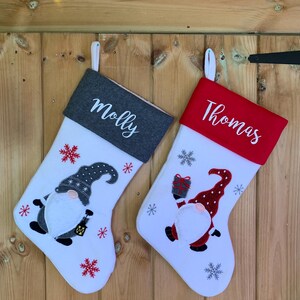 May include: Two white Christmas stockings with gnome embroidery. One stocking has a grey cuff with the name "Molly" embroidered on it. The other stocking has a red cuff with the name "Thomas" embroidered on it.