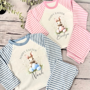 May include: Two pairs of pajamas with long sleeves and pants. The pajamas are white with blue and pink stripes on the sleeves and pants. The shirts have a bunny holding Easter eggs and the text "Happy Easter" and a name. One shirt says "Evelyn" and the other says "Noah".
