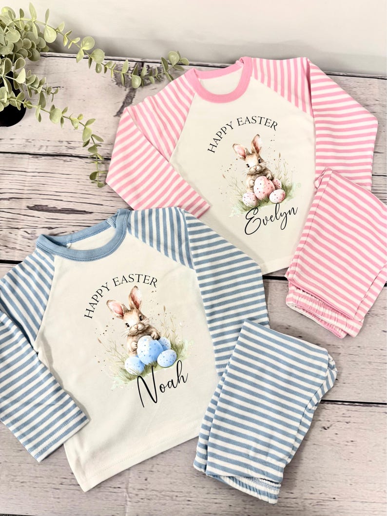 May include: Two pairs of pajamas with long sleeves and pants. The pajamas are white with blue and pink stripes on the sleeves and pants. The shirts have a bunny holding Easter eggs and the text "Happy Easter" and a name. One shirt says "Evelyn" and the other says "Noah".