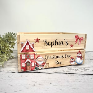 May include: A wooden Christmas Eve box with the name "Sophia's" on top. The box is decorated with a red house, Santa Claus, a reindeer, a penguin, and a snowman. The words "Christmas Eve Box" are also visible.