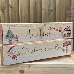 May include: A wooden Christmas Eve box with a snowman, Christmas tree, and Santa Claus design. The box is personalized with the name "Amelia's" and the text "Christmas Eve Box".