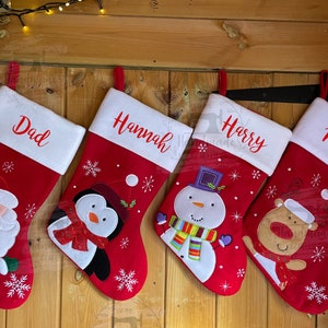 May include: Four red Christmas stockings with white cuffs hang on a wooden door. Each stocking is personalised with a name in red: Dad, Hannah, Harry, and Mum. Festive characters include Santa, a penguin, a snowman, and a reindeer.