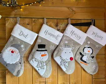 May include: Four personalized Christmas stockings with different embroidered designs. The stockings are gray with white trim and feature a snowman, reindeer, penguin, and Santa Claus. The stockings are labeled "Dad", "Hannah", "Harry", and "Mum".