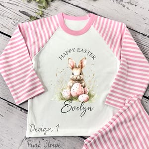 May include: A white and pink striped pajamas set with a long-sleeved shirt featuring a bunny rabbit holding Easter eggs and the text "Happy Easter" and "Evelyn". The shirt also has the text "Design 1 Pink Stripe".
