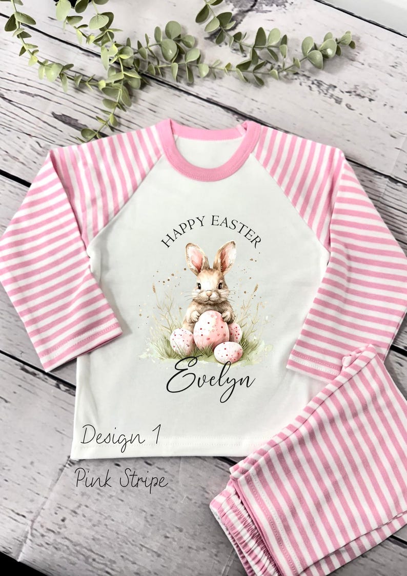 May include: A white and pink striped pajamas set with a long-sleeved shirt featuring a bunny rabbit holding Easter eggs and the text "Happy Easter" and "Evelyn". The shirt also has the text "Design 1 Pink Stripe".