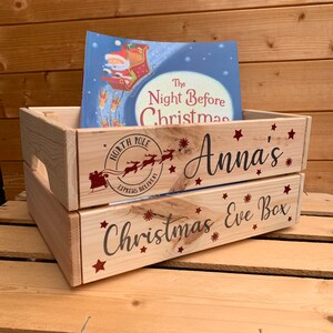 May include: A wooden crate with the words "Christmas Eve Box" and "Anna's" printed on it. The crate is decorated with red stars and snowflakes. A book titled "The Night Before Christmas" is sitting inside the crate.