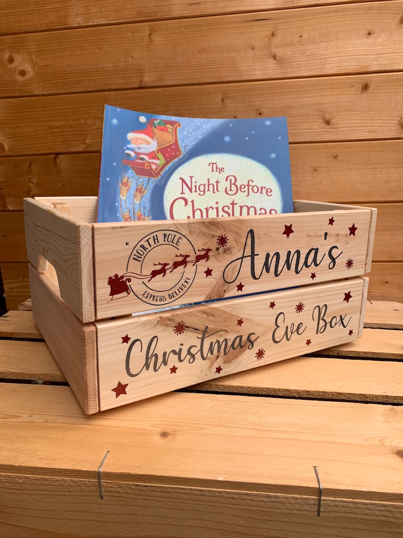 May include: A wooden crate with the words "Christmas Eve Box" and "Anna's" printed on it. The crate is decorated with red stars and snowflakes. A book titled "The Night Before Christmas" is sitting inside the crate.