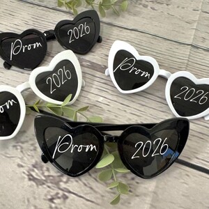 May include: Heart-shaped sunglasses in black and white, with the word "Prom" and the year "2026" printed on the lenses. The sunglasses are arranged on a wooden surface with green sprigs.