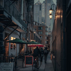May include: A narrow alleyway in a city with brick buildings on either side. A person rides a bicycle down the alleyway, passing by two other people walking. A sign for "The Tasting Room" is visible on the left side of the image.