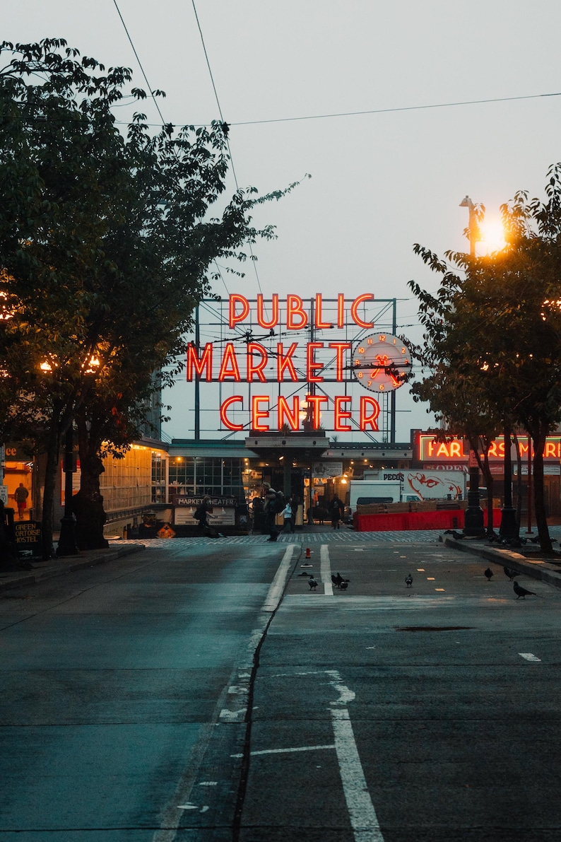 May include: Neon sign reading "PUBLIC MARKET CENTER" in red, above a street with trees and streetlights. A clock is integrated into the sign. The scene is at dusk, with a few people visible in the distance.