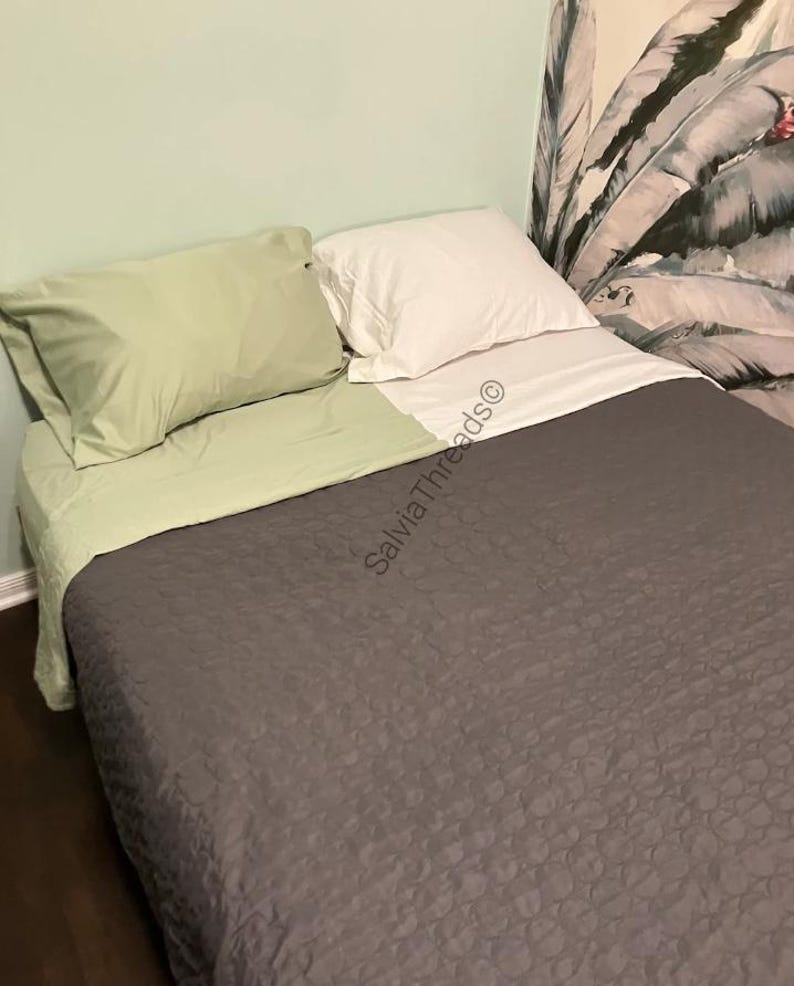 Puede incluir: Una cama con una colcha acolchada de color gris oscuro, una s&aacute;bana encimera de color verde claro, una funda de almohada verde claro a juego y una funda de almohada blanca. El texto SalviaThreads&copy; es visible en el centro.