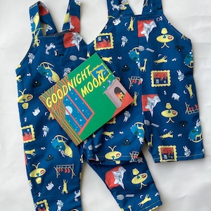 Puede incluir: Dos petos azules con un patrón repetitivo de ilustraciones del libro Goodnight Moon, que incluyen lámparas, sillas y cuadros. Un ejemplar del libro Goodnight Moon con cubierta verde y texto amarillo está colocado encima.