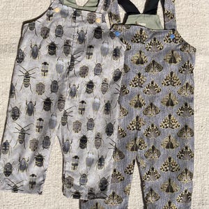 Puede incluir: Dos pares de petos de bebé. Un par presenta un estampado de varios insectos en tonos grises, negros y dorados sobre un fondo gris claro. El otro par tiene un estampado de polillas en colores similares. Ambos tienen tirantes ajustables.