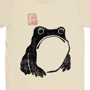 Könnte beinhalten: Ein beiges T-Shirt mit einem schwarz-weißen Bild einer Kröte und einem roten Stempel mit japanischen Schriftzeichen.
