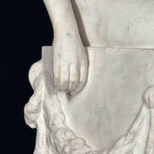May include: A white marble sculpture of a figure sitting on a block. The figure's right hand is resting on the block. The sculpture is detailed with intricate folds of fabric.
