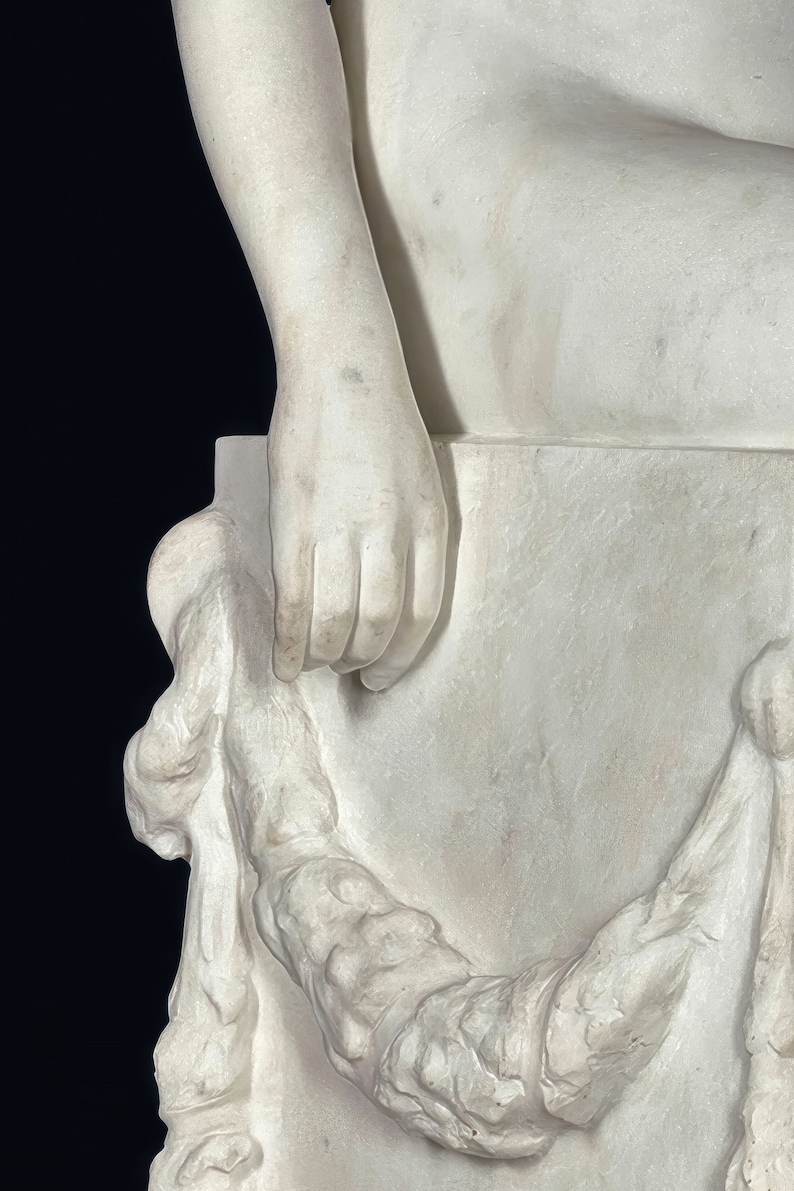 May include: A white marble sculpture of a figure sitting on a block. The figure's right hand is resting on the block. The sculpture is detailed with intricate folds of fabric.