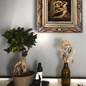 May include: A framed painting of a dramatic scene with three figures in a dark, fiery setting. The painting is in a gold ornate frame and is hanging on a light gray wall.