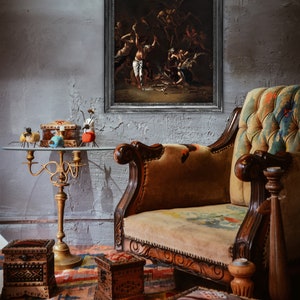 Pode incluir: Uma cena interior em estilo antigo com uma pintura emoldurada com um tema escuro e dram&aacute;tico. Um sof&aacute; vintage com detalhes em madeira ornamentados e uma almofada estampada fica ao lado de uma mesa com tampo de vidro com itens decorativos. Caixas de madeira e um tapete estampado completam a cena.