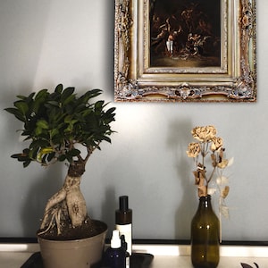 Pode incluir: Uma pintura ornamentada com moldura dourada est&aacute; pendurada numa parede cinzenta. Uma &aacute;rvore bonsai num vaso castanho, uma bandeja preta com garrafas e um vaso com flores secas est&atilde;o numa superf&iacute;cie branca. A pintura retrata uma cena escura com figuras.