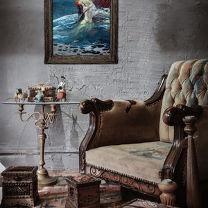 May include: A vintage-style, brown leather armchair with a floral patterned upholstery. The armchair is in a room with a framed painting of a mermaid and a table with decorative boxes.