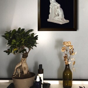 May include: A framed black and white photograph of a sculpture depicting two nude figures. The sculpture is on a pedestal with a garland of flowers. The photograph is hanging on a light gray wall.