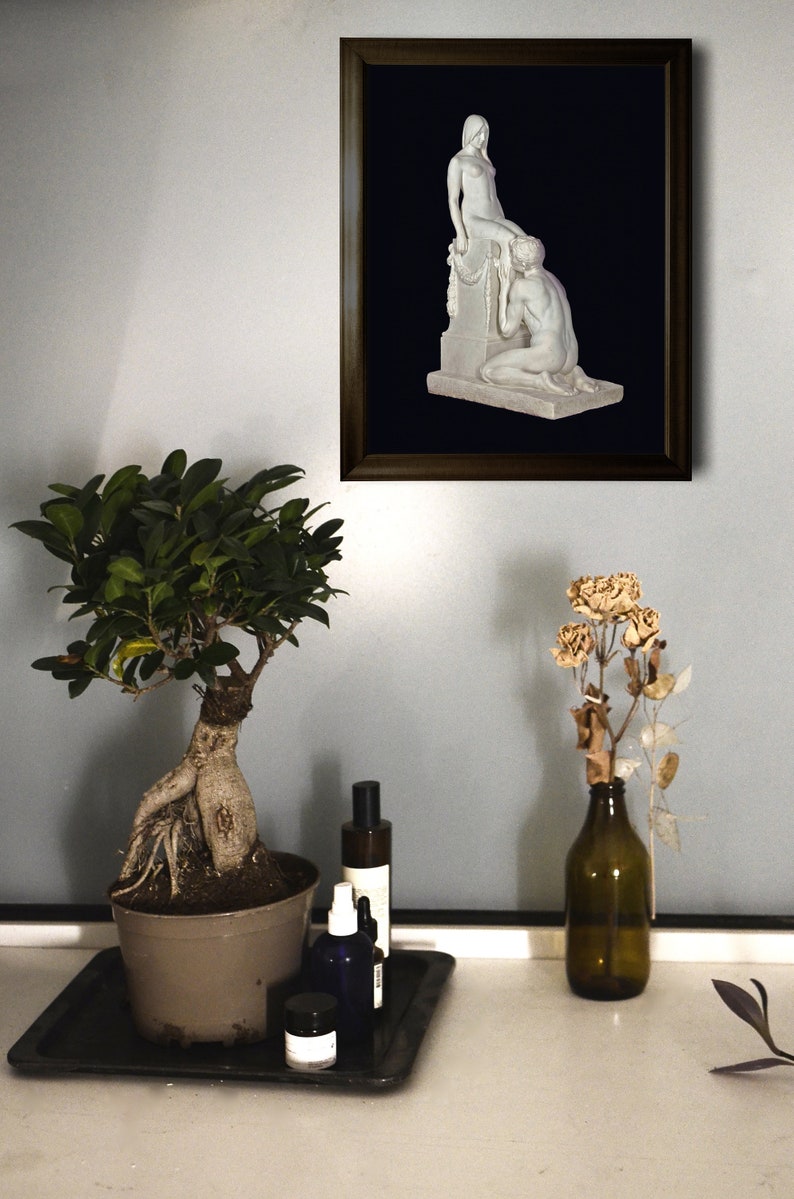 May include: A framed black and white photograph of a sculpture depicting two nude figures. The sculpture is on a pedestal with a garland of flowers. The photograph is hanging on a light gray wall.
