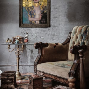 May include: A framed painting of a woman with long dark hair wearing a gold and blue robe. The painting is hanging on a gray brick wall above a brown velvet sofa. There are several small wooden boxes on a table in front of the sofa.