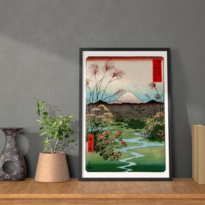 May include: A framed print of a Japanese woodblock print depicting Mount Fuji with a river and red flowers in the foreground. The print is in a black frame and has a white mat.