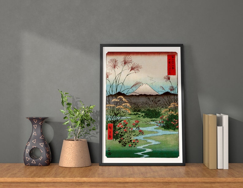 May include: A framed print of a Japanese woodblock print depicting Mount Fuji with a river and red flowers in the foreground. The print is in a black frame and has a white mat.