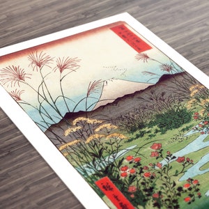 May include: A vintage Japanese woodblock print depicting Mount Fuji in the distance, with a winding river in the foreground. The print features a variety of plants and flowers, including red blossoms, green grasses, and yellow wildflowers. The print is framed in a white mat.