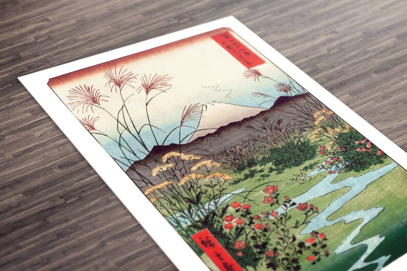 May include: A vintage Japanese woodblock print depicting Mount Fuji in the distance, with a winding river in the foreground. The print features a variety of plants and flowers, including red blossoms, green grasses, and yellow wildflowers. The print is framed in a white mat.
