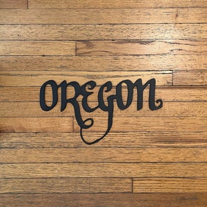 May include: Black metal sign with the word "Oregon" written in a stylized font. The sign is on a wooden floor.