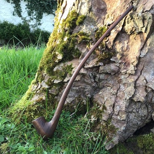 May include: A dark brown, long-stemmed wooden pipe with a curved design. The pipe is set against a tree trunk covered in moss. The background features green grass and foliage. The pipe is likely for smoking.