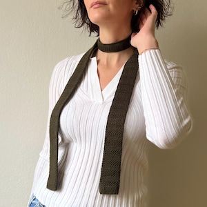 May include: Olive green knitted necktie. The tie is a solid color and has a textured appearance. The tie is worn around the neck and hangs down the front of a white long-sleeved shirt.