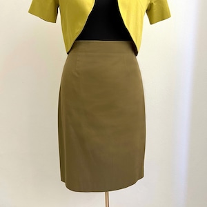 Può includere: Gonna a tubino verde oliva abbinata a una giacca chartreuse a maniche corte. La gonna è lunga fino al ginocchio e ha un design classico. La giacca è aperta sul davanti, rivelando un top nero. L'outfit è esposto su un manichino.