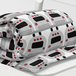 May include: A gray and white fleece blanket with a repeating pattern of cartoon subway trains. The blanket is rolled up and placed on a white chair.