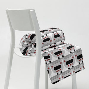 May include: A white chair with a gray and white blanket with a repeating pattern of gray and black buses with red headlights. The blanket is rolled up on the chair.