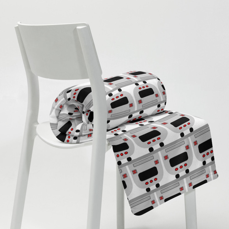 May include: A white chair with a gray and white blanket with a repeating pattern of gray and black buses with red headlights. The blanket is rolled up on the chair.