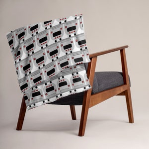 May include: A white throw blanket with a repeating pattern of gray and black buses. The blanket is draped over a wooden chair with a gray cushion.