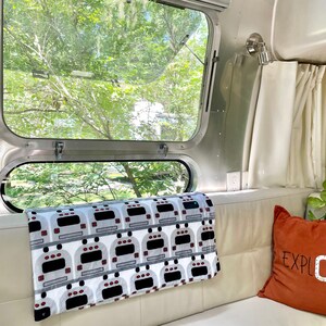 May include: A white couch with a gray and white throw blanket with a pattern of Airstream trailers. A rust-colored pillow with the word "EXPLORE" in white lettering is on the couch. A window with a view of green trees is behind the couch.