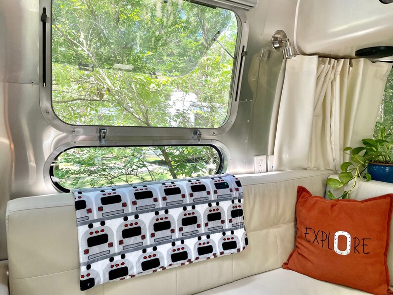 May include: A white couch with a gray and white throw blanket with a pattern of Airstream trailers. A rust-colored pillow with the word "EXPLORE" in white lettering is on the couch. A window with a view of green trees is behind the couch.