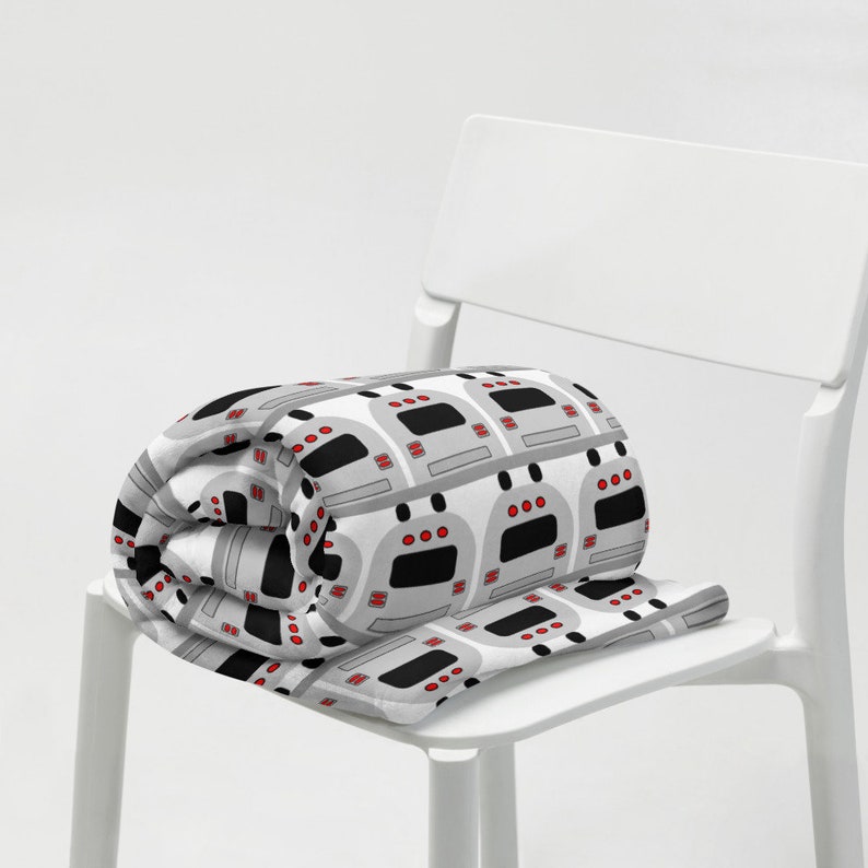 May include: A gray and white fleece blanket with a repeating pattern of cartoon subway trains. The blanket is rolled up and placed on a white chair.