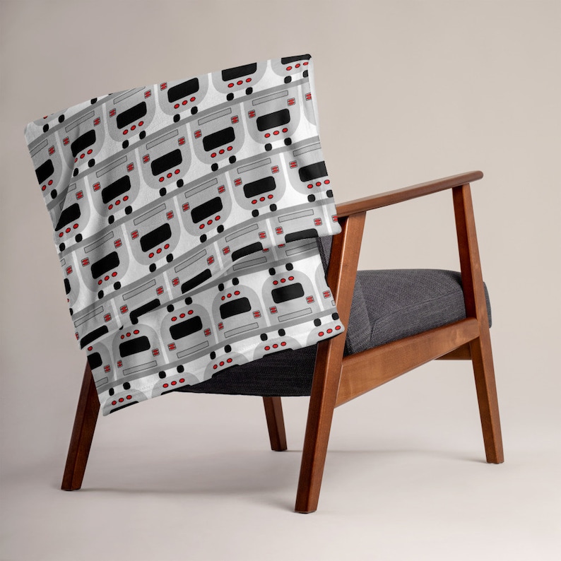 May include: A white throw blanket with a repeating pattern of gray subway trains. The blanket is draped over a wooden chair with a gray cushion.