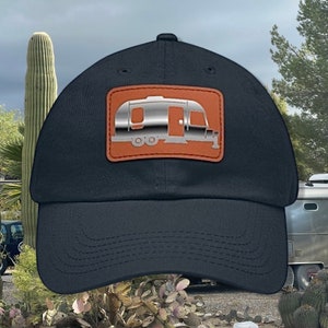 May include: A black baseball cap with a brown leather patch featuring a silver camper silhouette.
