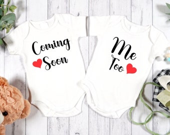 May include: Two white baby onesies with black text and red hearts. The onesie on the left says "Coming Soon" and the onesie on the right says "Me Too".