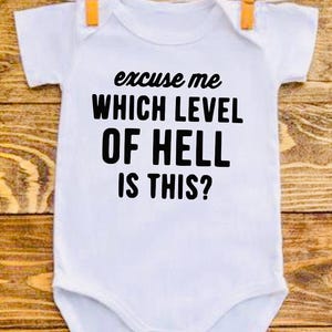 May include: A white short-sleeved baby bodysuit featuring the black printed text "excuse me WHICH LEVEL OF HELL IS THIS?" centred on the front. The garment has a round neckline and leg openings, designed for infants with a simple, clean aesthetic.