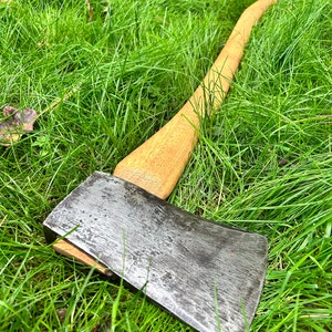 May include: A vintage axe with a wooden handle and a steel blade, resting on a bed of green grass. The axe head shows signs of wear, suggesting it has been used. The wooden handle is a light brown color.