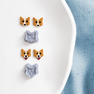 Puede incluir: Una colección de accesorios con temática de corgi, que incluye dos pares de pendientes de botón con cabeza de corgi y dos amuletos grises con forma de cara de corgi. Los pendientes presentan caras de corgi naranjas y blancas con ojos negros y lenguas rojas. Los artículos están colocados en un plato blanco.