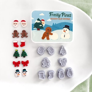 May include: A set of Frosty Pines earring clay cutters in a rectangular box with a winter scene. Included are six pairs of small, colourful holiday-themed clay shapes: Santa faces, gingerbread people, stockings, trees, bows, and snowmen.